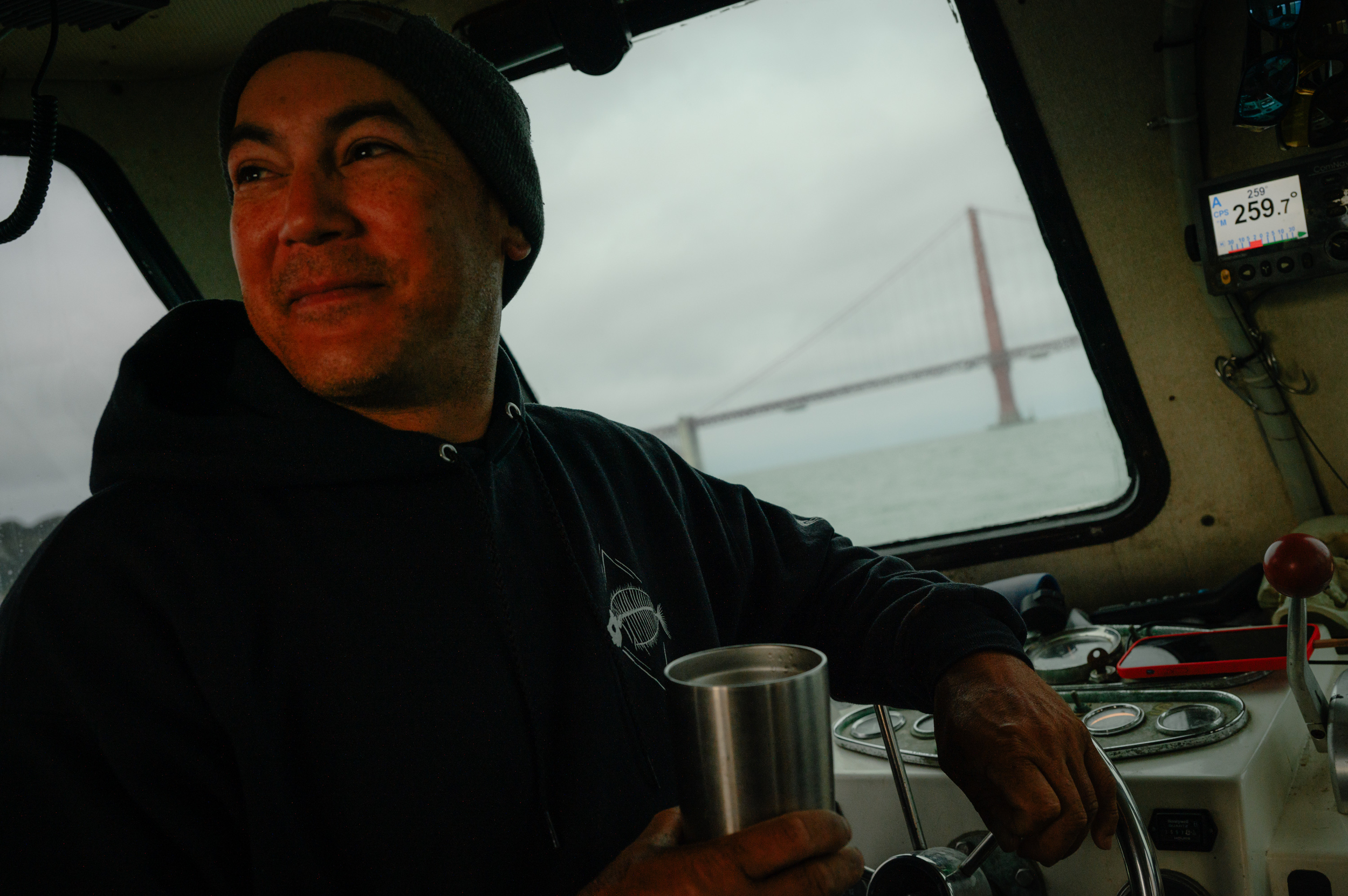 A man smiles holding a cup of coffee and leaning on the steering wheel of a boat. Through the window over his shoulder is the Golden Gate Bridge.