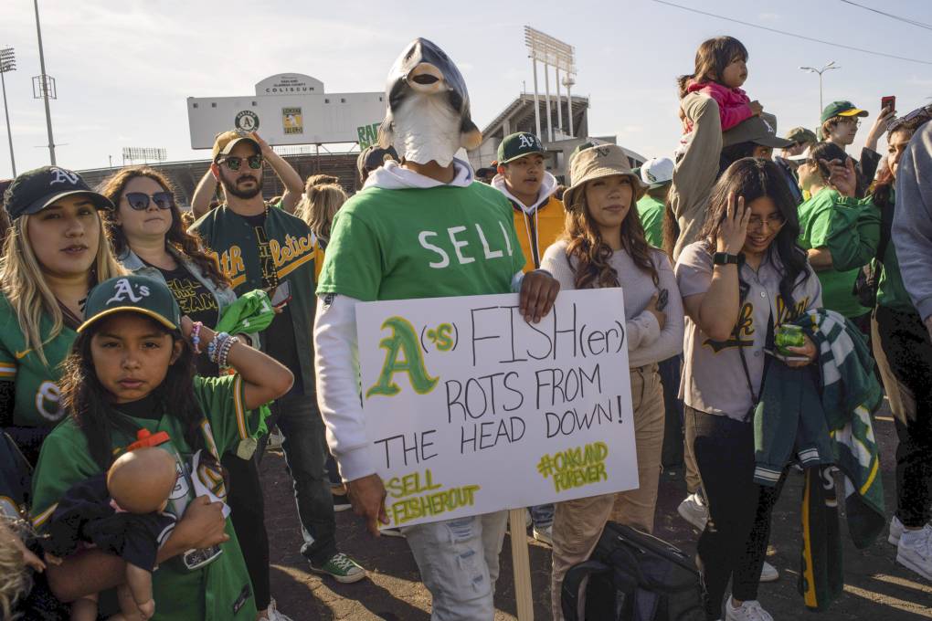 A crowd of people wearing Oakland A's clothing stand together in a parking lot. The person in the middle wears a plastic fish mask and holds a sign reading "A's Fish(er) rots from the head down!"