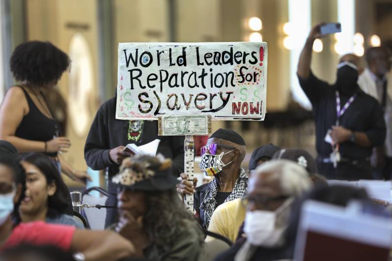 A man wearing a face mask holds a sign that reads "World Leaders! Reparations for Slavery Now!" in a crowd of people.