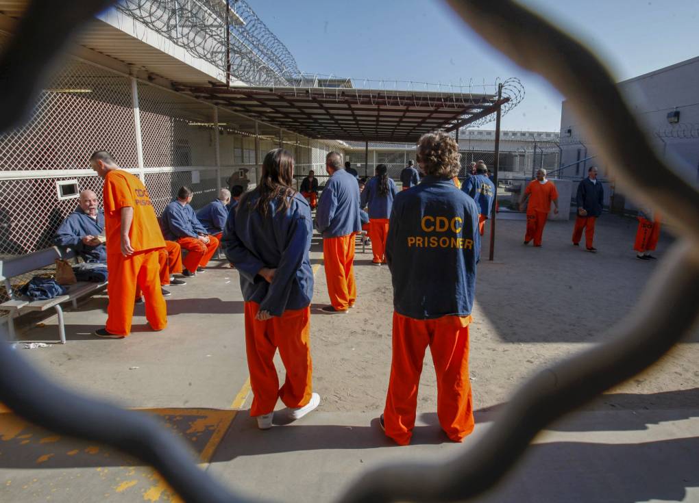 Men dressed in blue long-sleeved shirts and bright orange pants stand and walk in an open outdoor area surrounded by fencing and barbed wire.