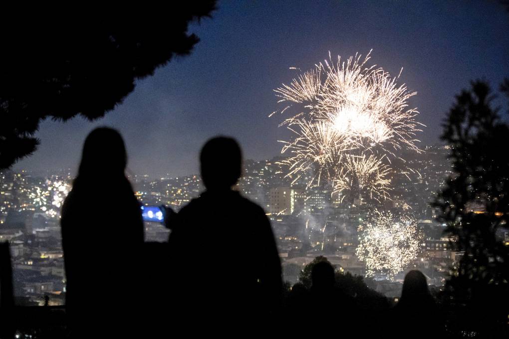 The silhouettes of two or more people are thrown into relief by the explosions of fireworks in the distance over a city setting.
