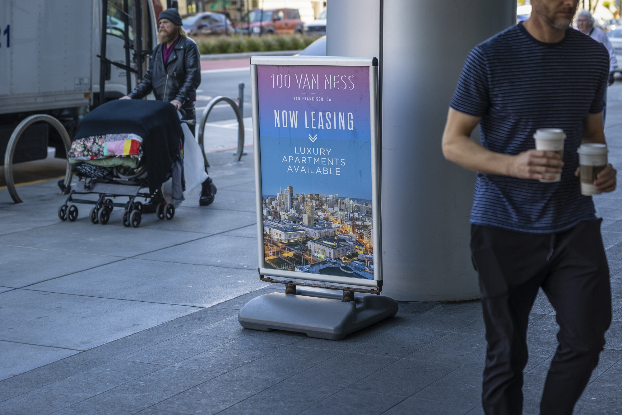 People pass a sign on a city sidewalk advertising luxury apartments.