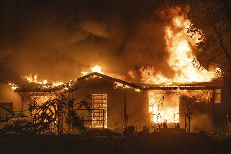 In a nighttime photo lit dramatically by orange flames, a single-story home burns brightly, with the photo taken from the end of a driveway or yard.