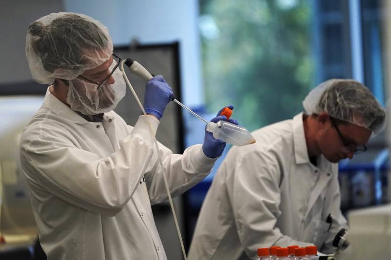 Two white men are wearing white lab coats, hair covers, masks and blue surgical gloves. The man on the left is standing, holding a device with a long needle-like end going into a specimen container.
