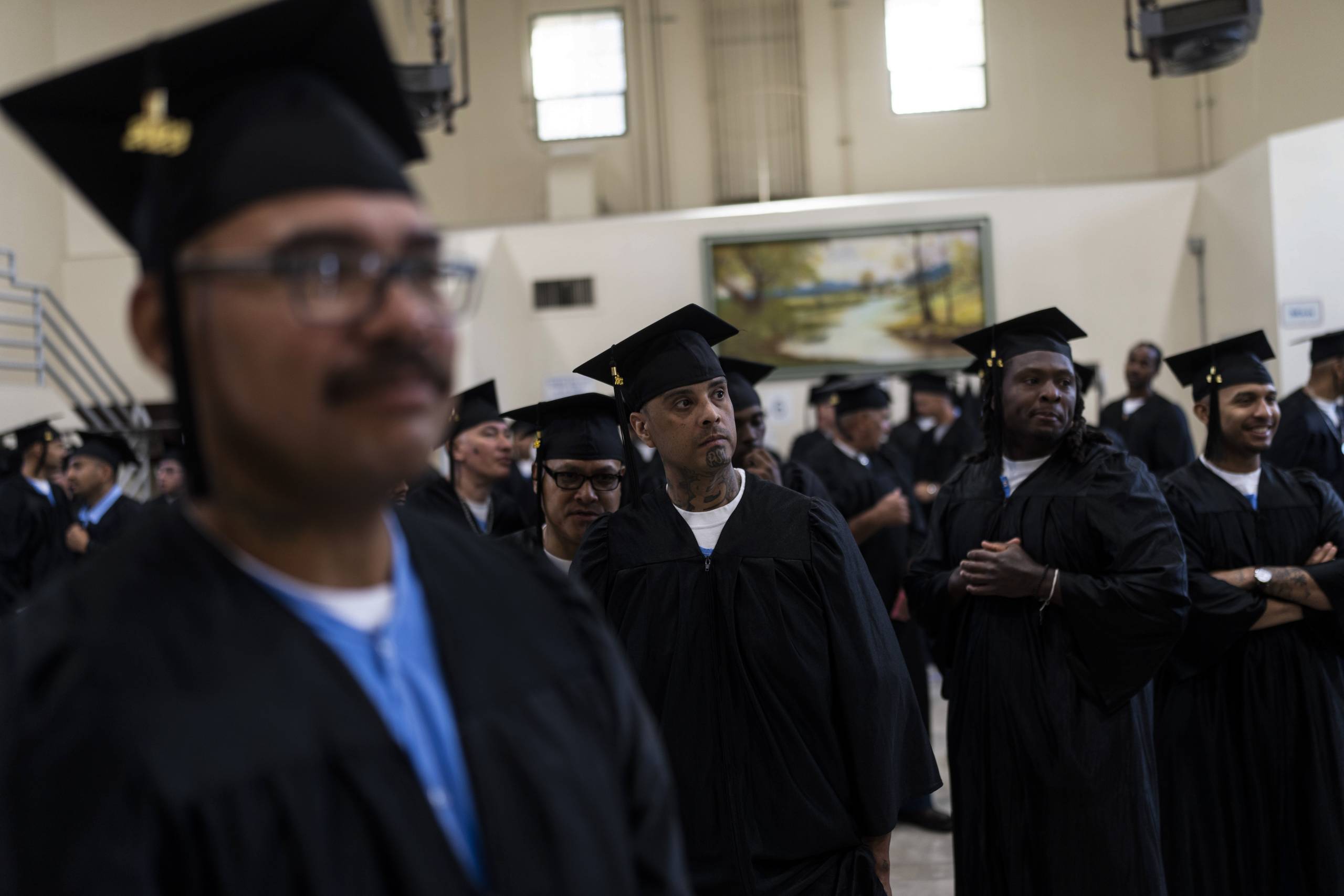 A group of men of mixed ages, wearing graduation caps and gowns on top of blue prison gowns, gather in a large room.