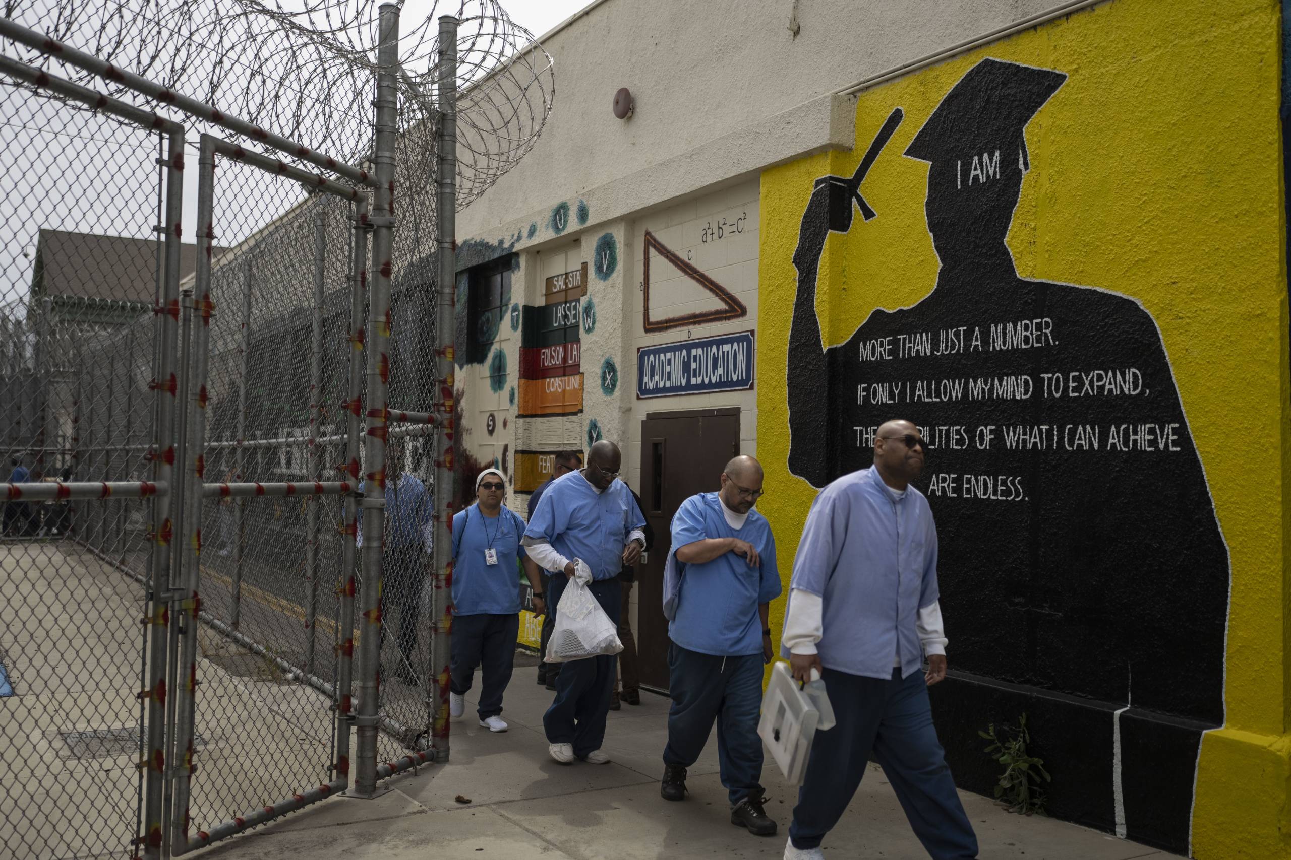 A group of men, wearing blue prison shirts, walk out of a fenced yard, past a mural of a person in a gown, that says: 'More than a number.'