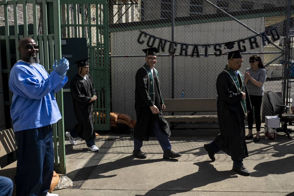 Three young men in black graduation gowns walk through a prison courtyard, as a man in a blue gown looks on.