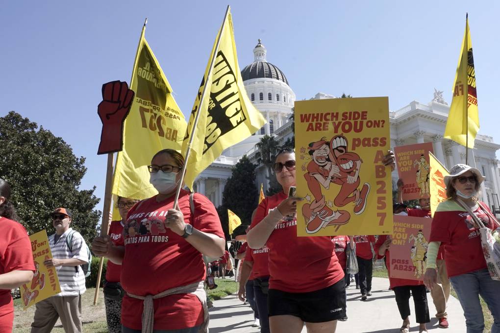 A large group of protestors wearing red T-shirts march around California's Capitol building in Sacramento. Many are carrying yellow signs with red writing. One sign reads, "Which Side Are You On? Pass AB257."