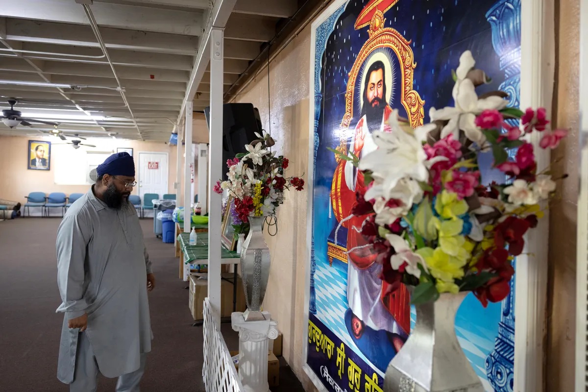 A bearded Indian man with glasses, a dark blue turban priest, and a long, light-blue tunic stands before a wall-size, colorful graphic illustration of another bearded Indian man.