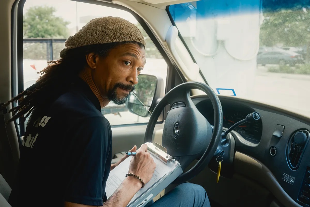 A man with long dreadlocks and a tan hat sits inside his vehicle as he writes on a piece of paper that's attached to a clipboard. A parking lot of cars is seen in the background, along with trees. The key is in the vehicle's ignition.