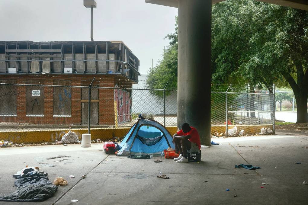 A man sits on a milk crate outside of his small, blue tent. A barbed-wire fence surrounds a building in the background. He appears to have set up a small camp under an overpass. A few trees are pictured in the background as well. The sky is gray. Trash is littered around his area.