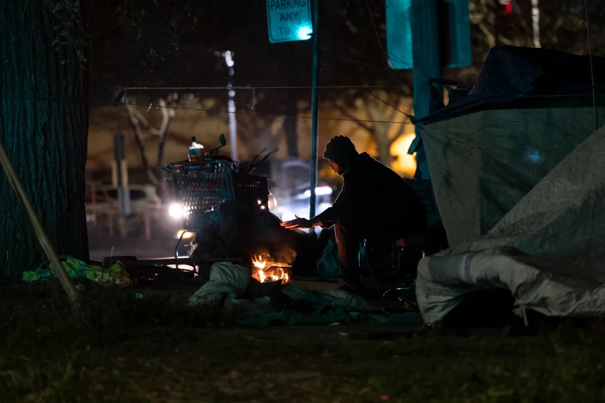 A man is seen outside of his makeshift tent, with a shopping cart full of his belongings, warming his hands by a small fire. It's nighttime. A "No Parking Anytime" sign in the background.