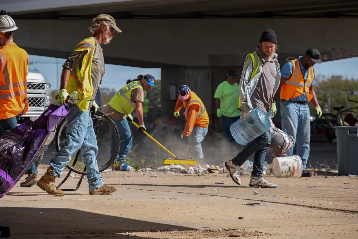 City workers in yellow and orange reflective vests work out in the hot Houston heat cleaning up an encampment. A man is pictured using a push-broom in the background.