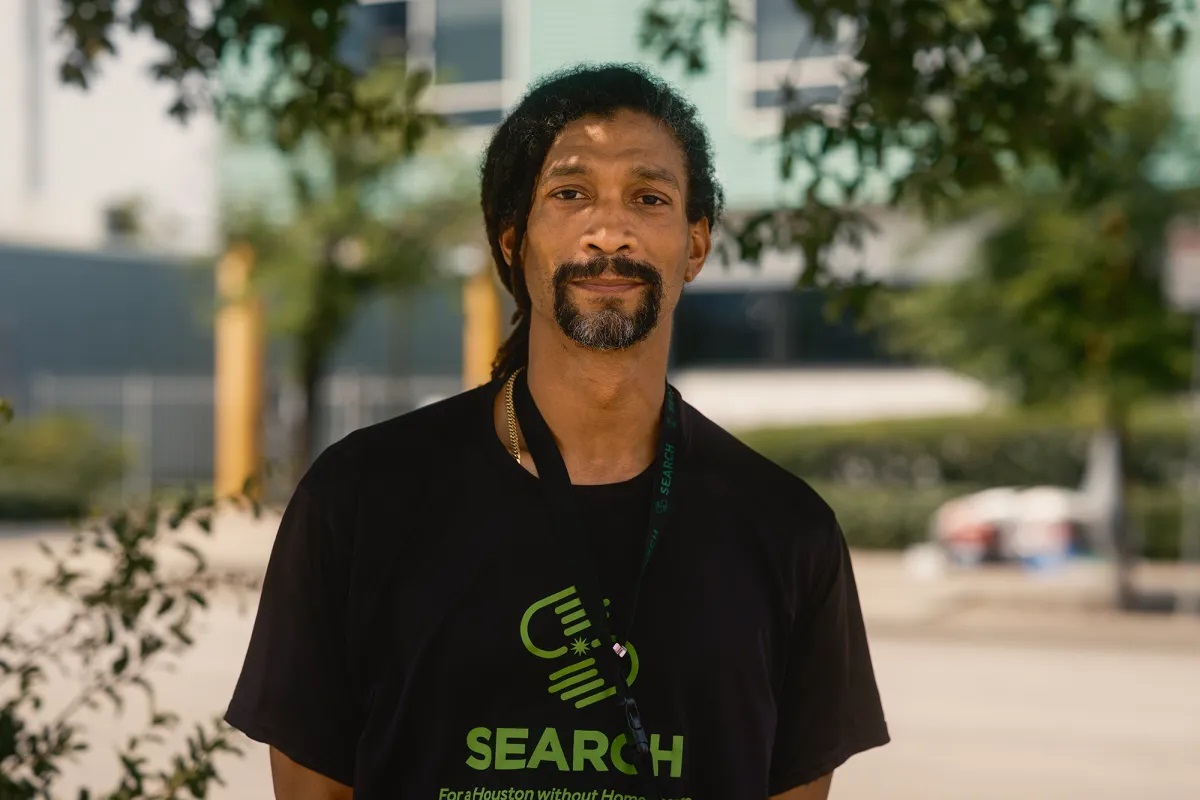 A man with calm expression and a friendly smile poses for a portrait. He has a T-shirt on that reads, "Search."