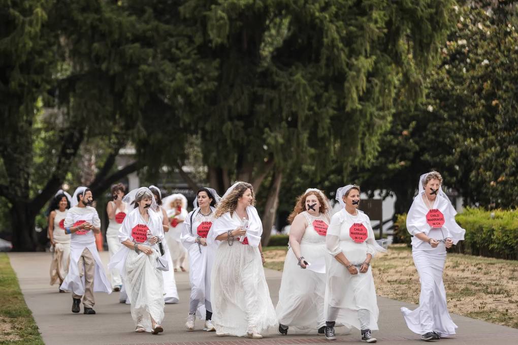 Protestors in white wedding gowns and lacey veils walk toward California's Capitol Building. Some have black tape over their mouths in an X shape. Each individual also wears a red stop sign on their chest that reads, "Stop Child Marriage in CA."