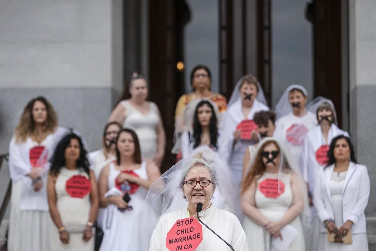 Many women wearing wedding dresses and veils are seen on the steps of California's Capitol Building. One older woman with gray hair and glasses, is speaking from a podium. All the individuals in the photo wear a red stop sign on their chest that reads, "Stop Child Marriage in CA."