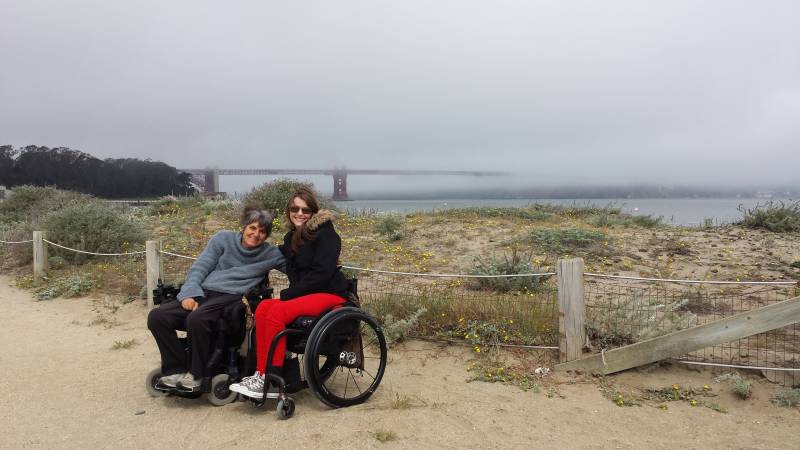 Two women with lighter skin and dark hair using wheelchairs are seen smiling against a backdrop of a foggy day covering up a red bridge also known as the Golden Gate Bridge. 
