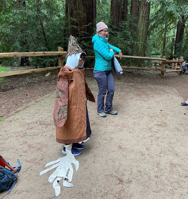 A child dressing in a redwood tree costumes stands next to a woman in a bright blue sweater. In the background, a redwood forest is visible.