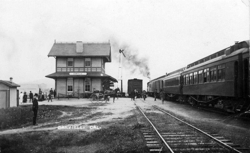 Black and white image of a train station. People mill about near the building. A steam engine is on the tracks, and a horse and carriage sits outside the building.