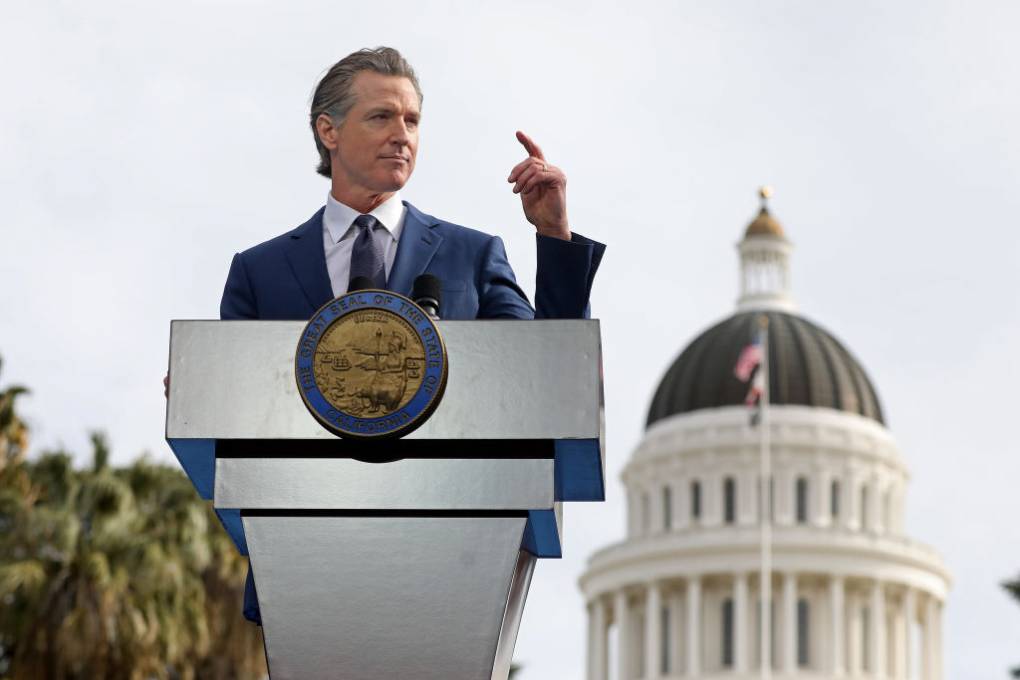 Gavin Newsom stands at a speaking podium, with the dome of the capitol building in the background.