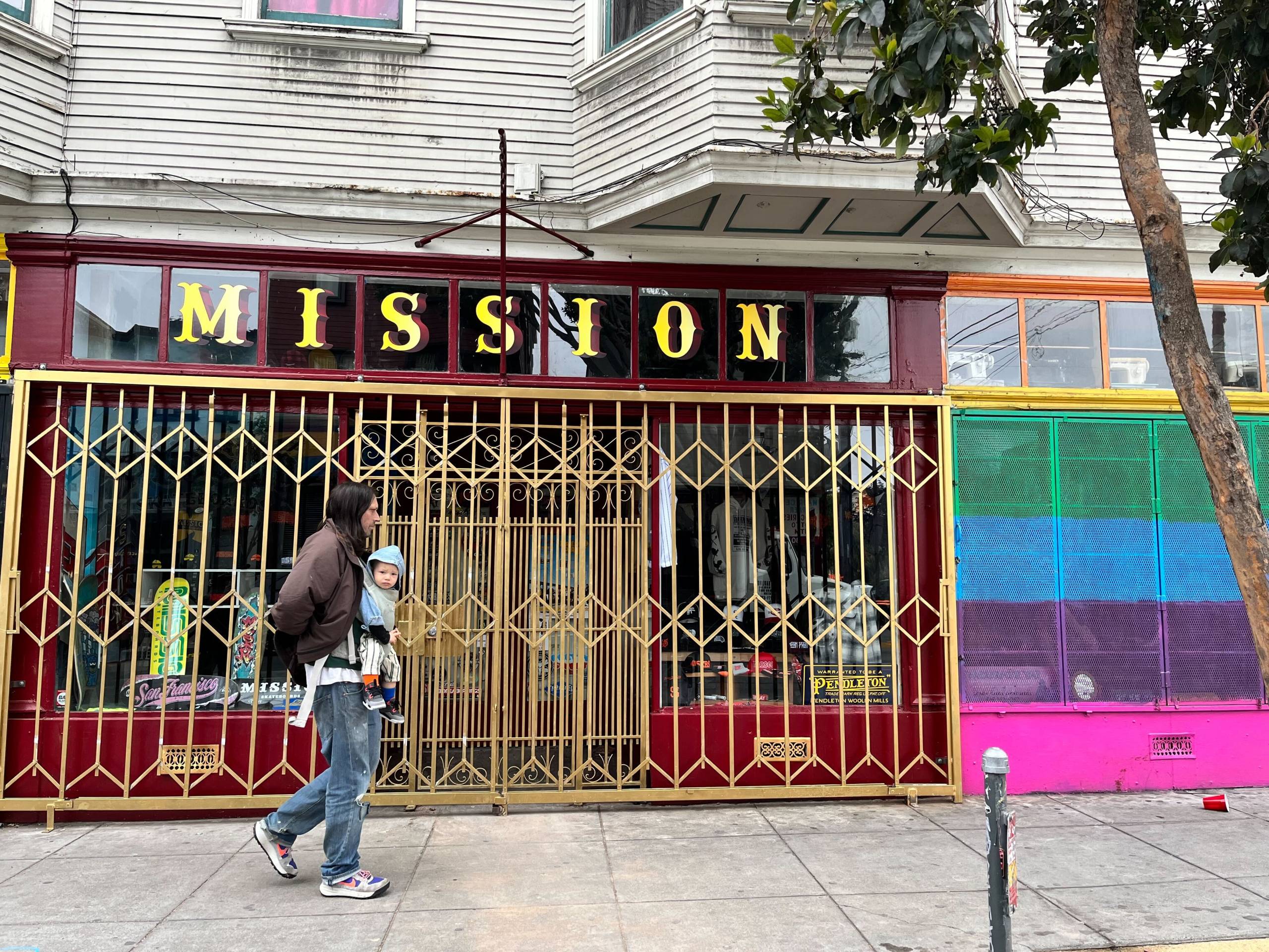 A closed storefront with "Mission" written over the main window, and a man with a toddler walking by.