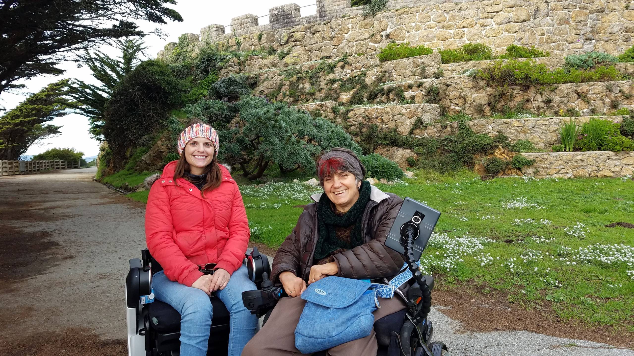 Two women with lighter skin and dark hair using wheelchairs are next to each other smiling against a backdrop of a fort, and trees. The woman on the left is wearing a red jacket. And the woman on the right is wearing a brown jacket holding a blue bag in her lap.