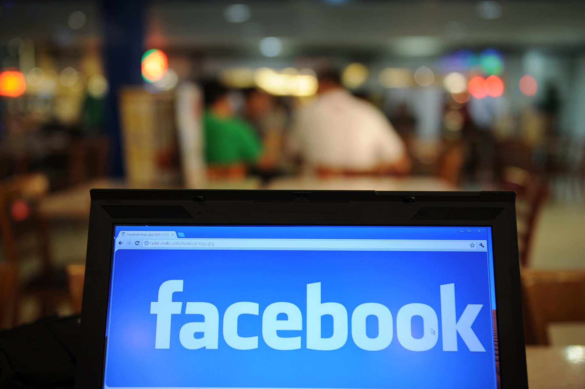 A computer tablet screen glows with a blue and white social media logo for the company Facebook. People are blurred in the background at a cafe setting.