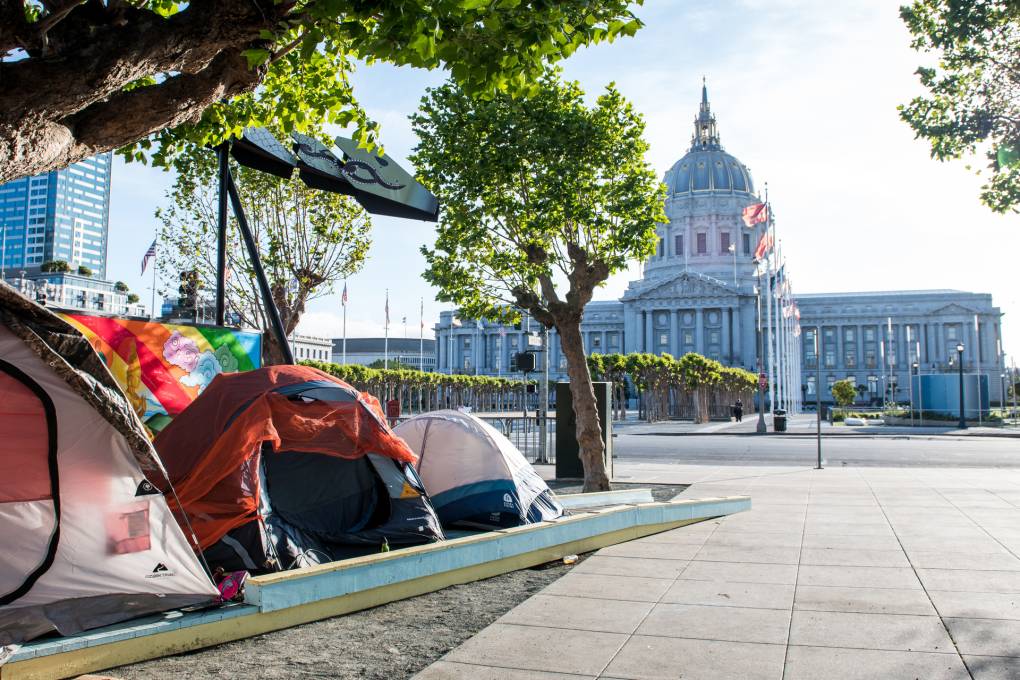 Three tents line a public street with San Francisco's City Hall building shining as the sun comes up in the early morning hours. Rows of trees line a path toward the city building.