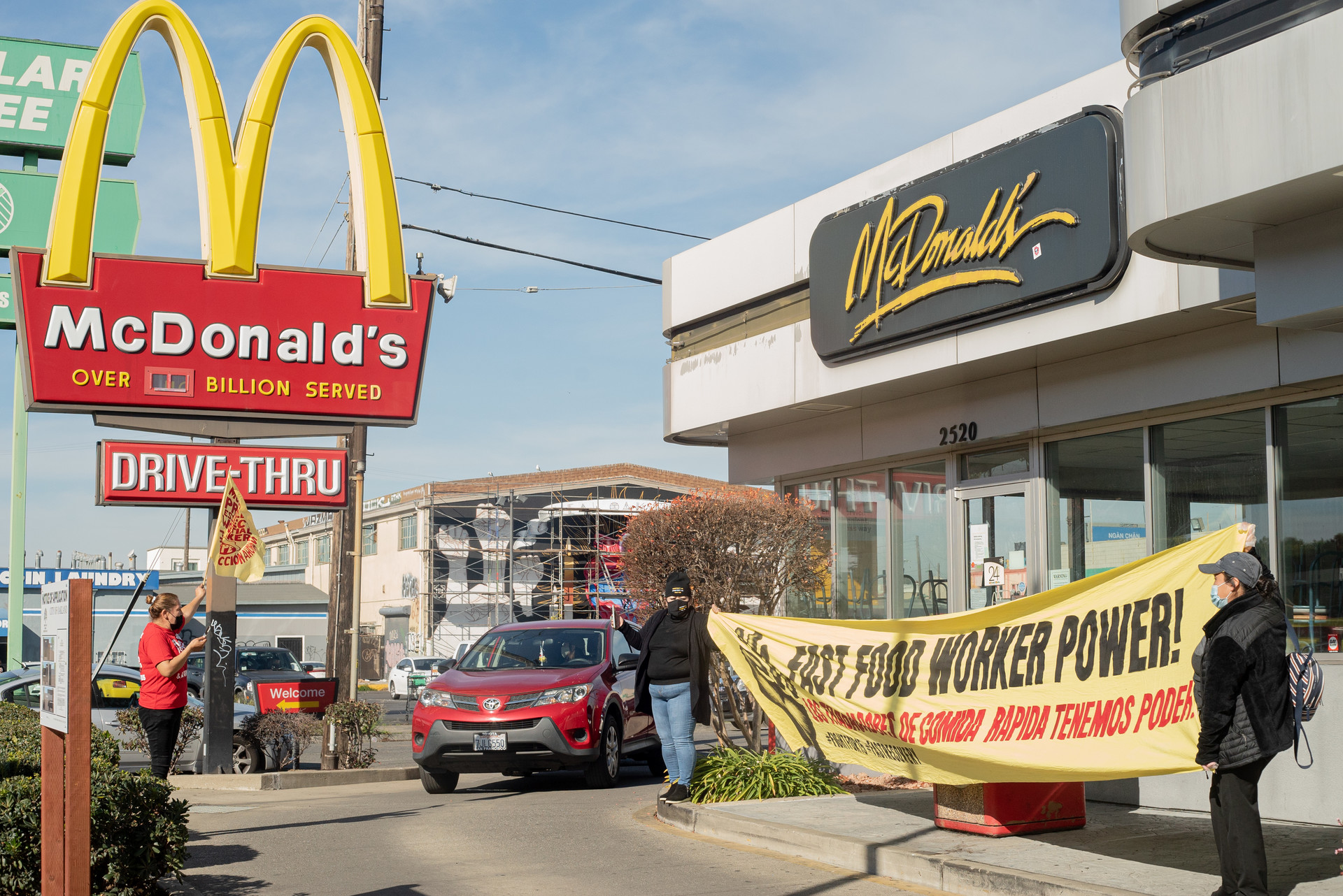 A McDonald's drive-thru is pictured where several protestors are seen holding a large yellow banner with black and red writing that reads, "Fast Food Worker Power!" A maroon SUV is pulling out of the drive-thru.