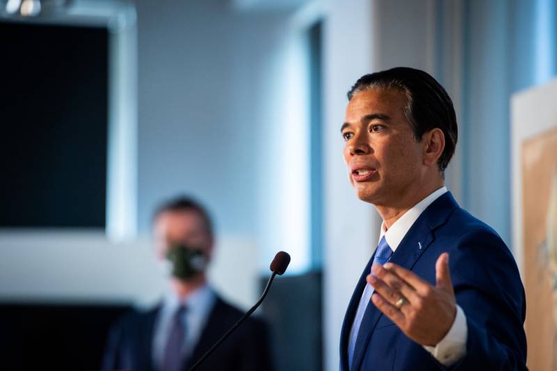 A man in a navy blue suit and tie speaks from a podium with one hand extended out as he wears a serious expression. His black hair has little gray. Gov. Gavin Newsom is seen standing in the background, blurred.