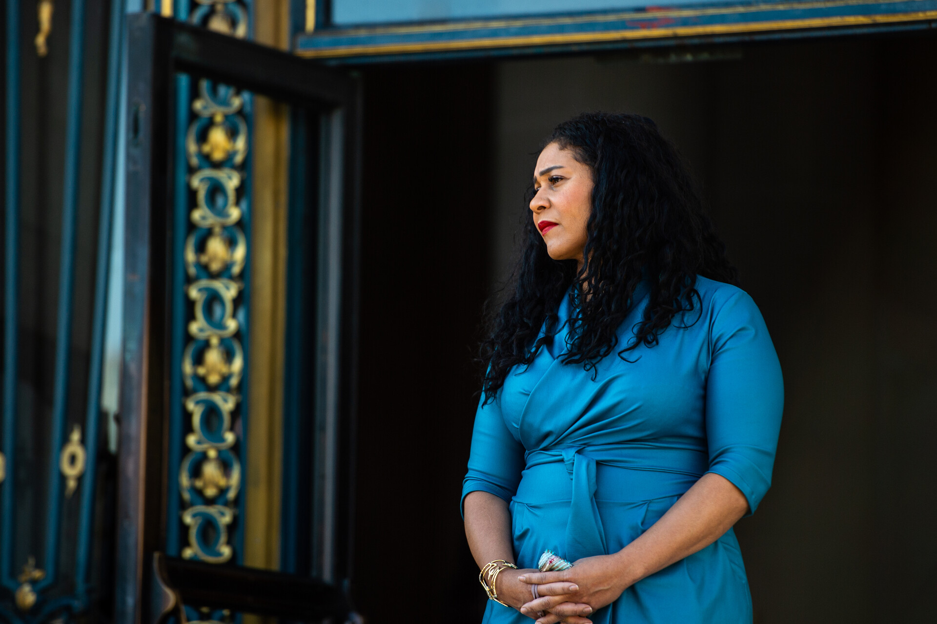 A woman with brown hair and a light blue dress stands with her hands folded in front of her. She is looking toward the left. She is standing in front of San Francisco's City Hall building listening to a speech.