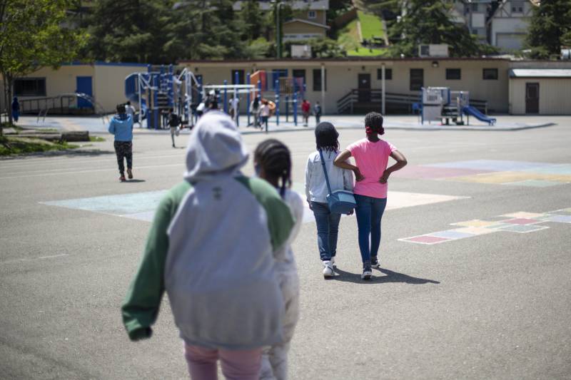A number of young people walk and run across a tarmac in the direction of a playground.