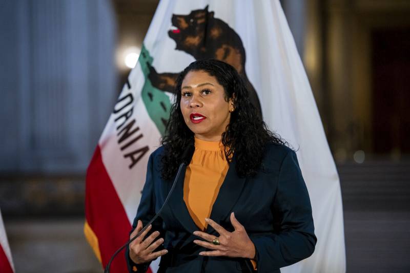 A Black woman with shoulder-length black, curly hair and bright red lipstick, wearing a blue blazer and an ochre blouse, gestures as she speaks in front of a flag of California inside what appears to be a large room.