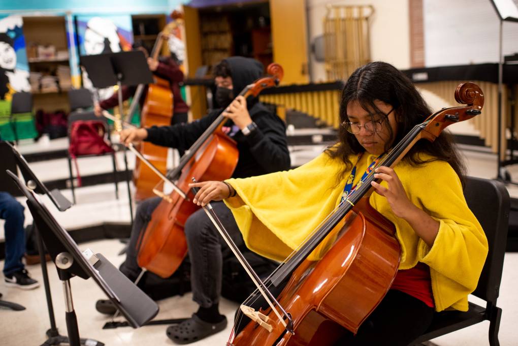A young teenage girl, with dark hair and glasses, plays a cello in a music classroom, with other students in the background.