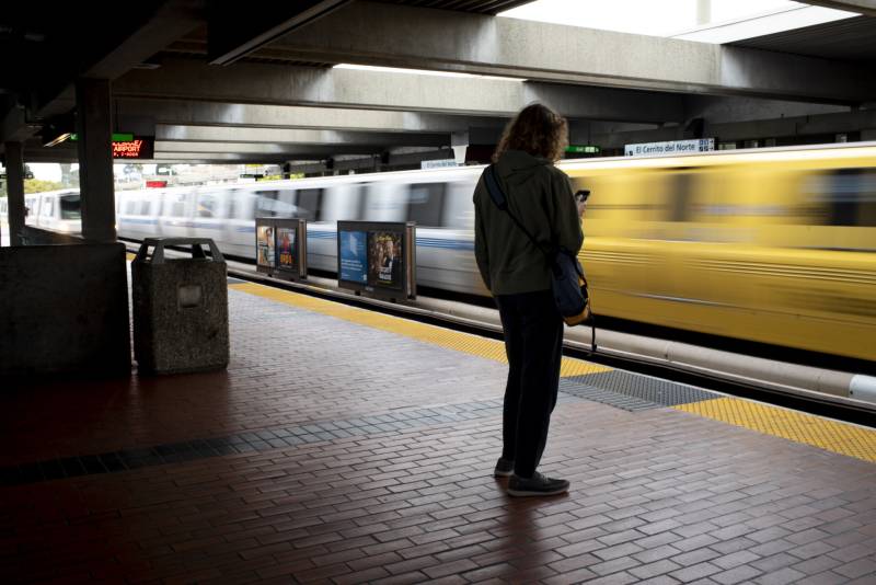 A person stands on a BART platform as a train arrives.