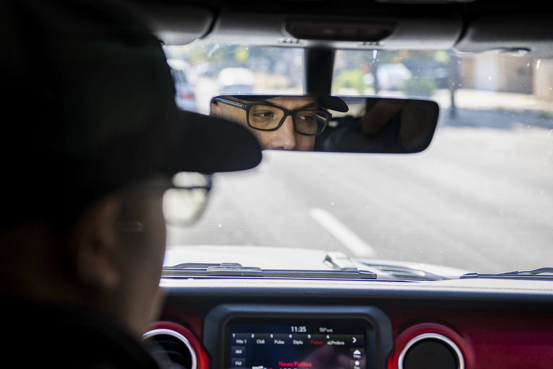 A photo taken from the backseat of a vehicle from behind the driver's side. Blurry in the foreground, and in focus in the rearview mirror, we see a light-skinned man in a black baseball cap driving and looking to the right.