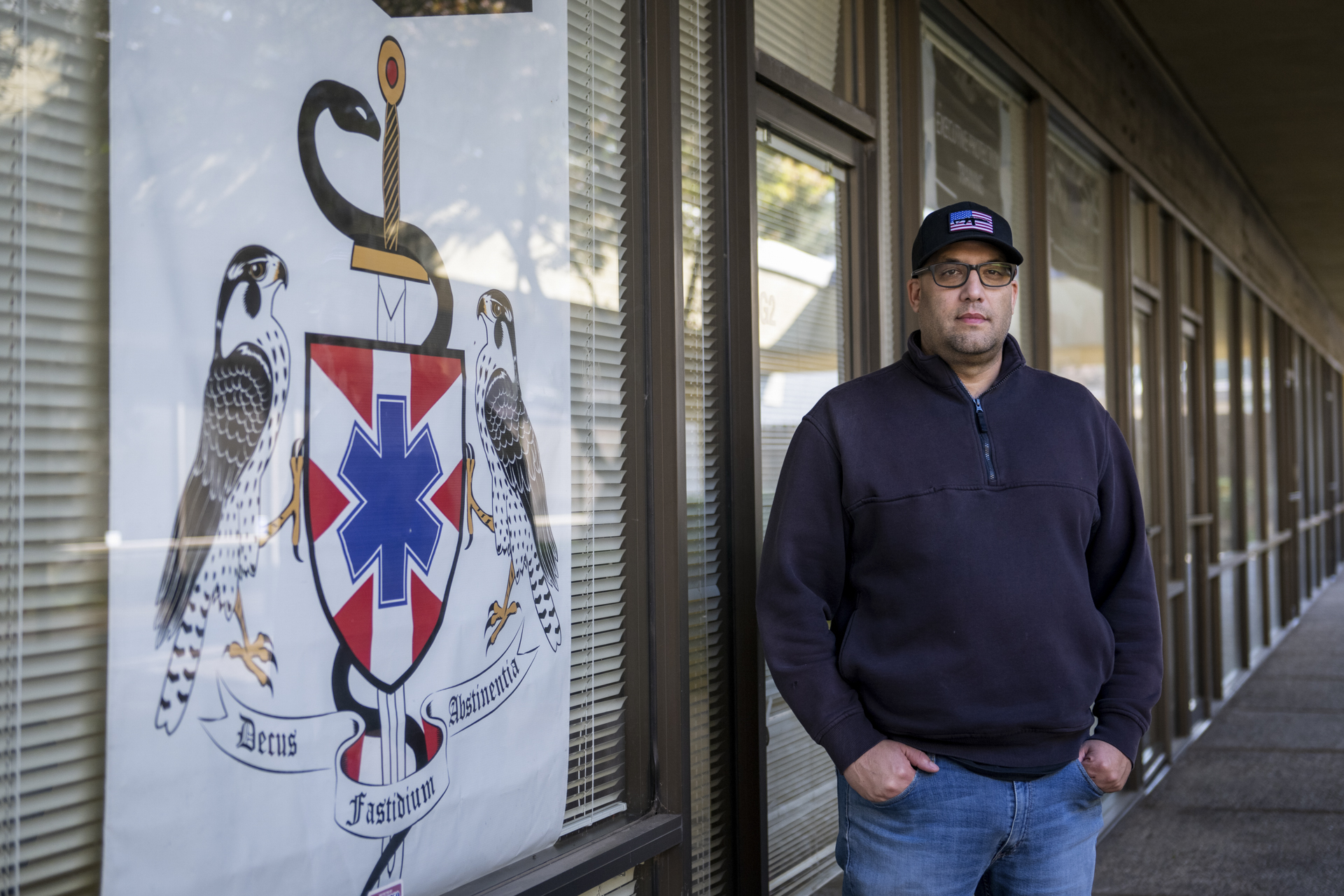 A white man looks seriously at the camera standing in front of a storefront at a strip mall next to a banner showing an insignia featuring a snake and two falcons. The man wears a black hat with an American flag, glasses, a dark fleece, and blue jeans.