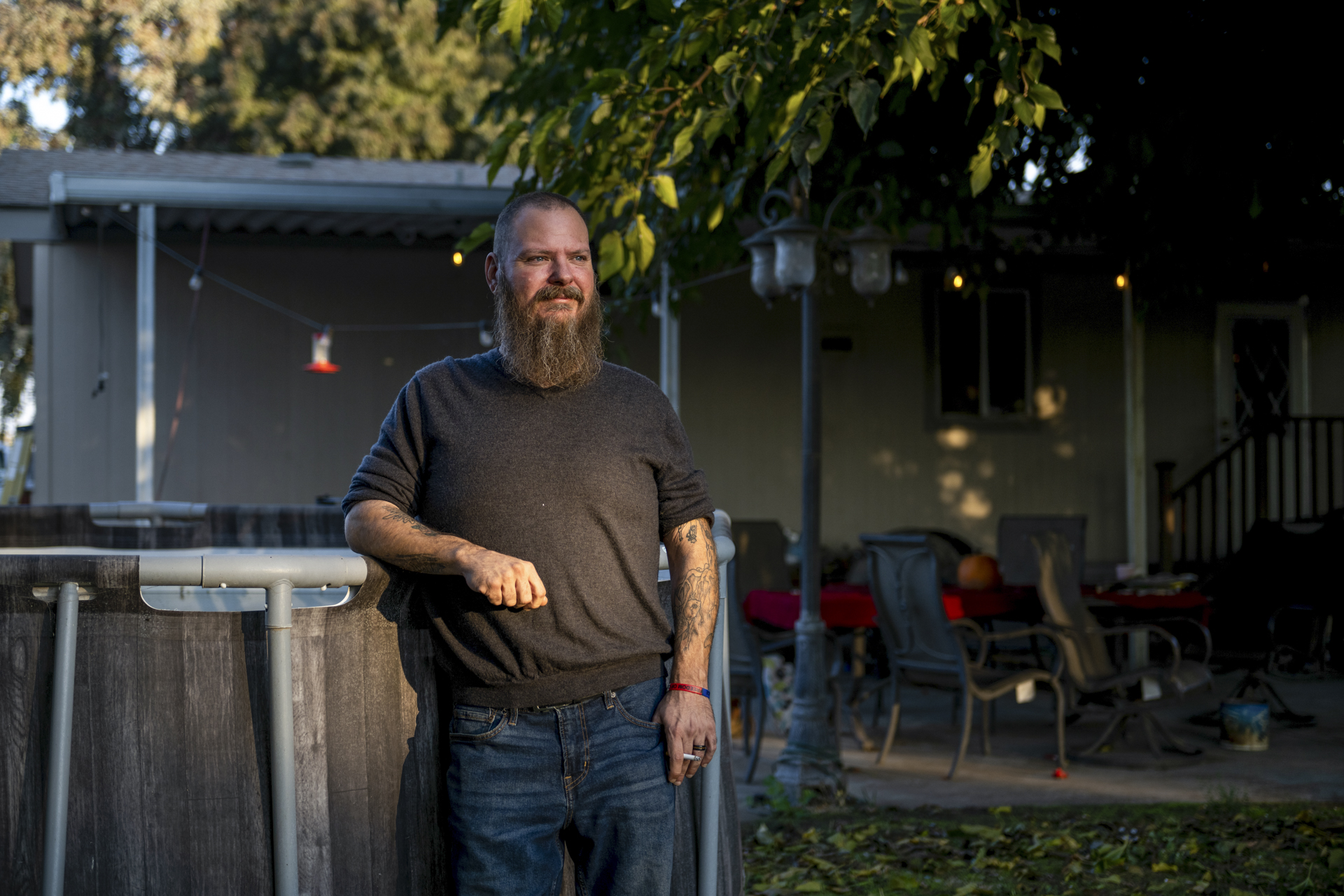 A white man with a long graying beard and shaved head leans against the edge of an above-ground swimming pool in the backyard of a home. He has tattoos on his arms and holds a cigarette in his left hands, and he wears baggy dark blue jeans and a dark gray sweatshirt.