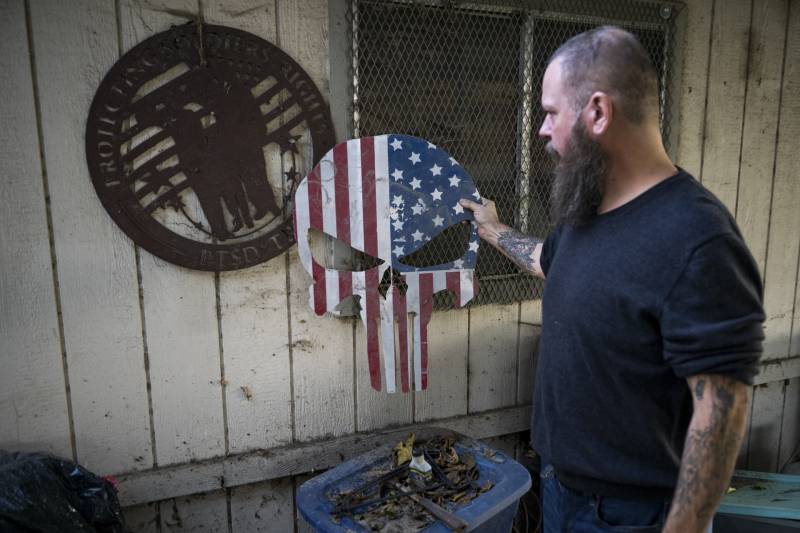 A man with a long beard and shaved head holds a skull-shaped carving painted in the style of the American flag outdoors.