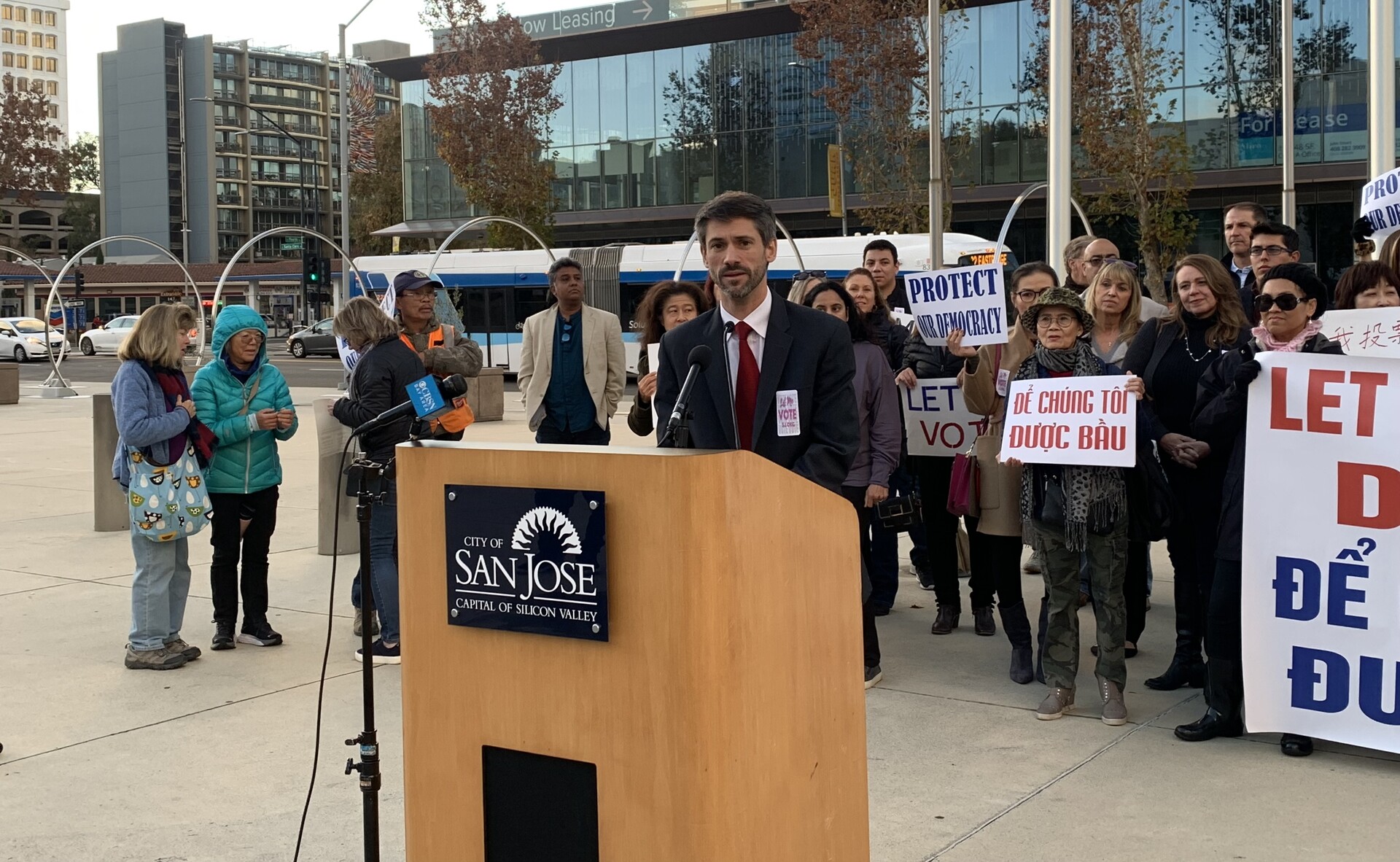 A man with a beard wearing a suit and tie stands behind a podium labeled, "San José Capital of Silicon Valley." He is surrounded by people holding various signs. Some read, "Protect our Democracy."