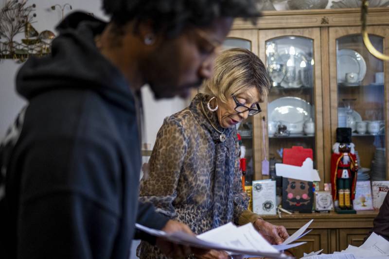 An older Black woman wearing a leopard print shirt goes through papers alongside a man doing the same. Behind the woman dishes are displayed on shelves and next to a nutcracker and other Christmas decorations.