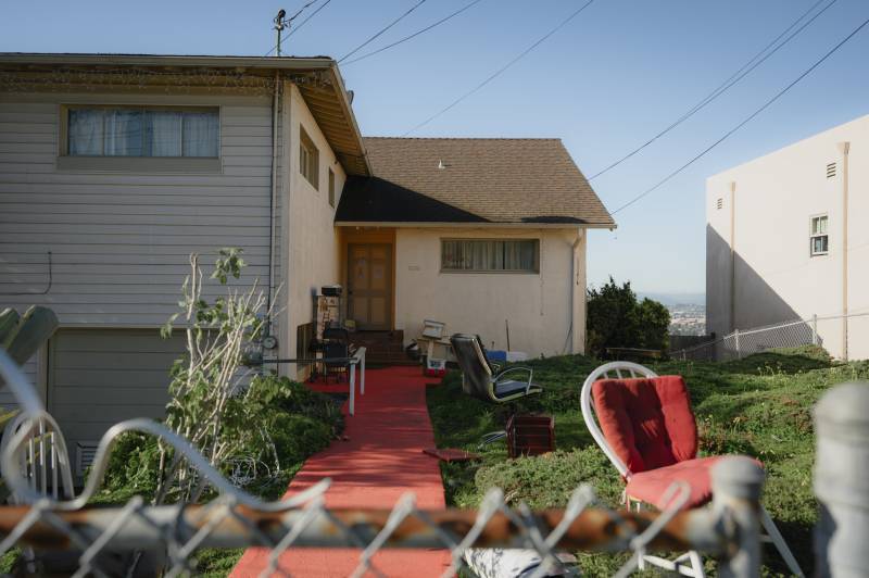 A front entrance to a home seen from outside the chainlink fence surrounding it. The front lawn has chairs and other belongings on it.