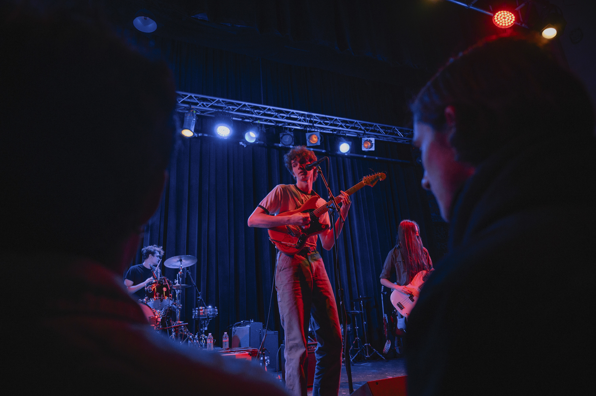 A young person plays a guitar while singing into a microphone alongside accompanying musicians playing the drums and the bass guitar under neon red and blue lights.