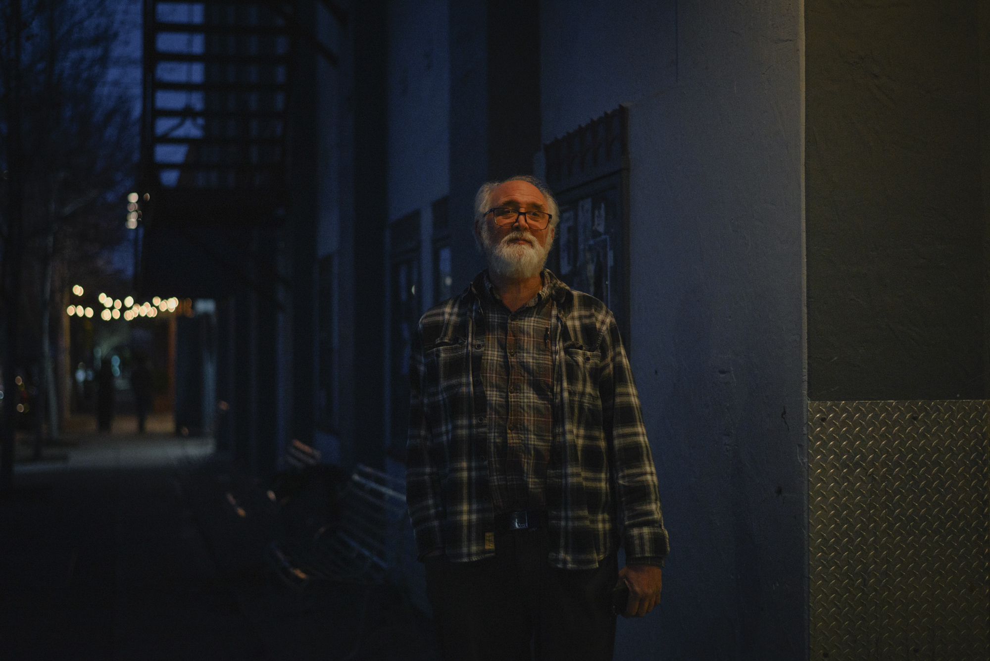A partly bald man with a white beard stands on a darkened sidewalk and looks at the camera.