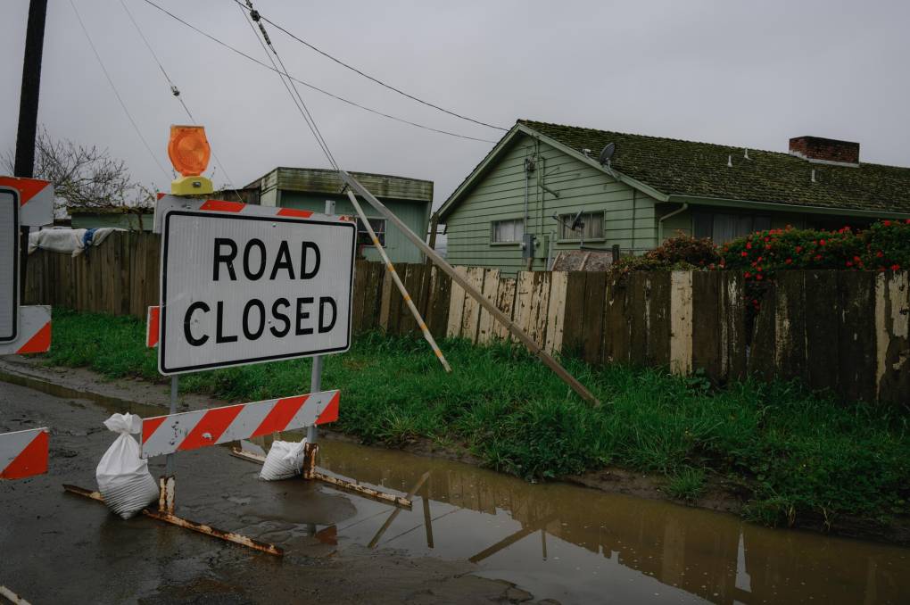 An orange and white construction sign reads, "Road closed," next to flooded streets and dilapidated wooden fences. A green, wooden home is seen in the background.