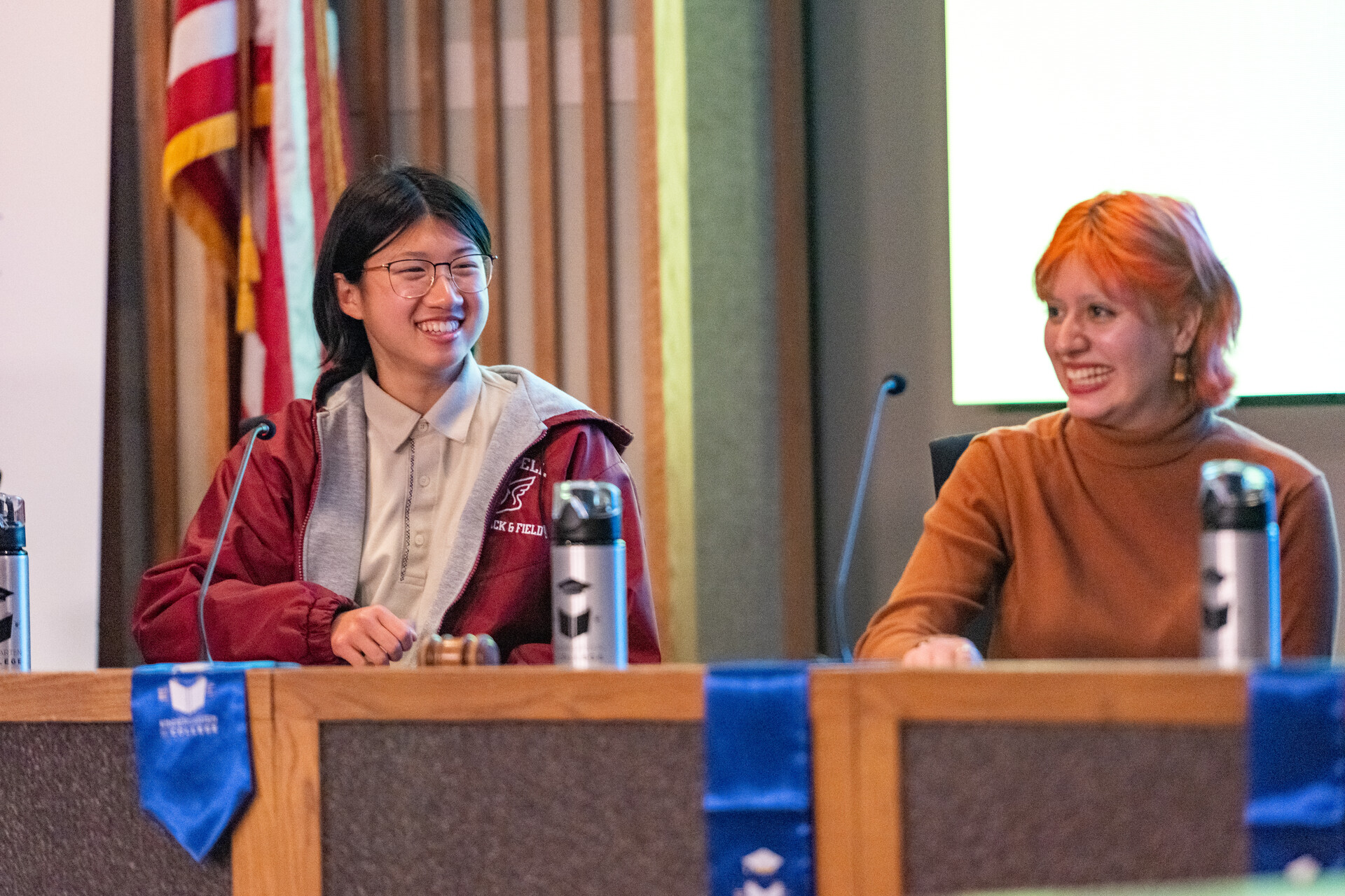 An Asian youth and a white youth smile with dyed orange hair sit behind a podium and smile at an unseen audience.