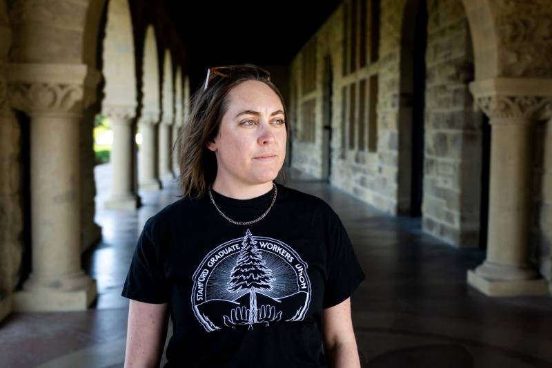 A woman with long, brown hair stares off into the distance. She stands in an outdoor hallway of a building with chunky pillars. She wears a black T-shirt that reads, "Stanford Graduate Workers Union."