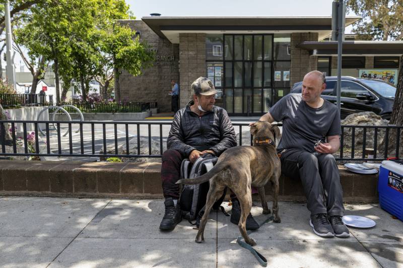 Two middle-aged white men sit on a bench in front of a building as one of them pets a dog.