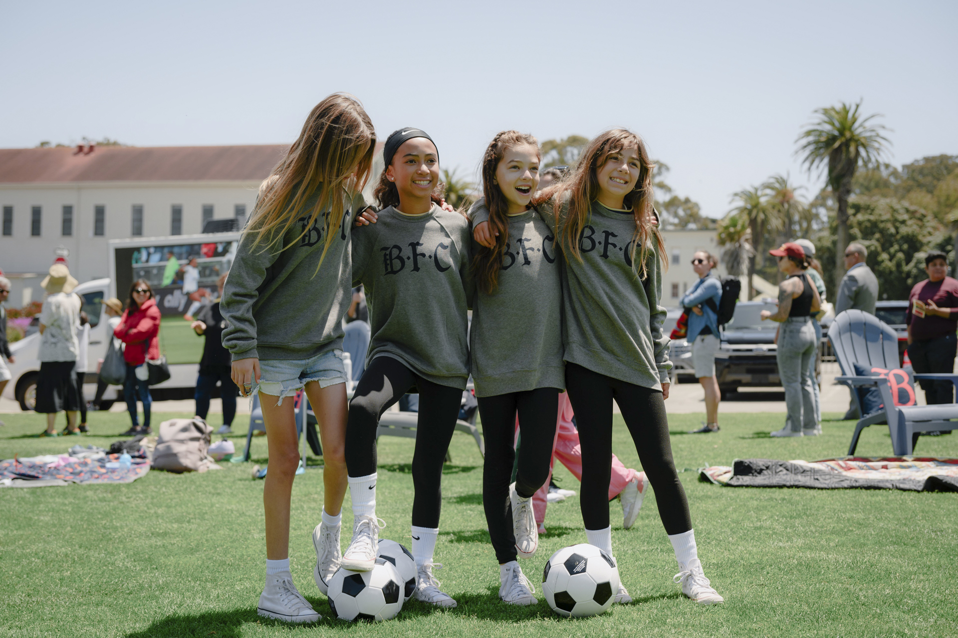 Four girls pose for a photo with two soccer balls and people in the background.