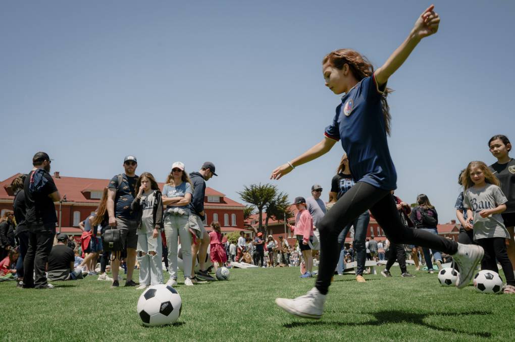 A girl with long hair about to kick a soccer ball while people look on at a lawn on a blue day.